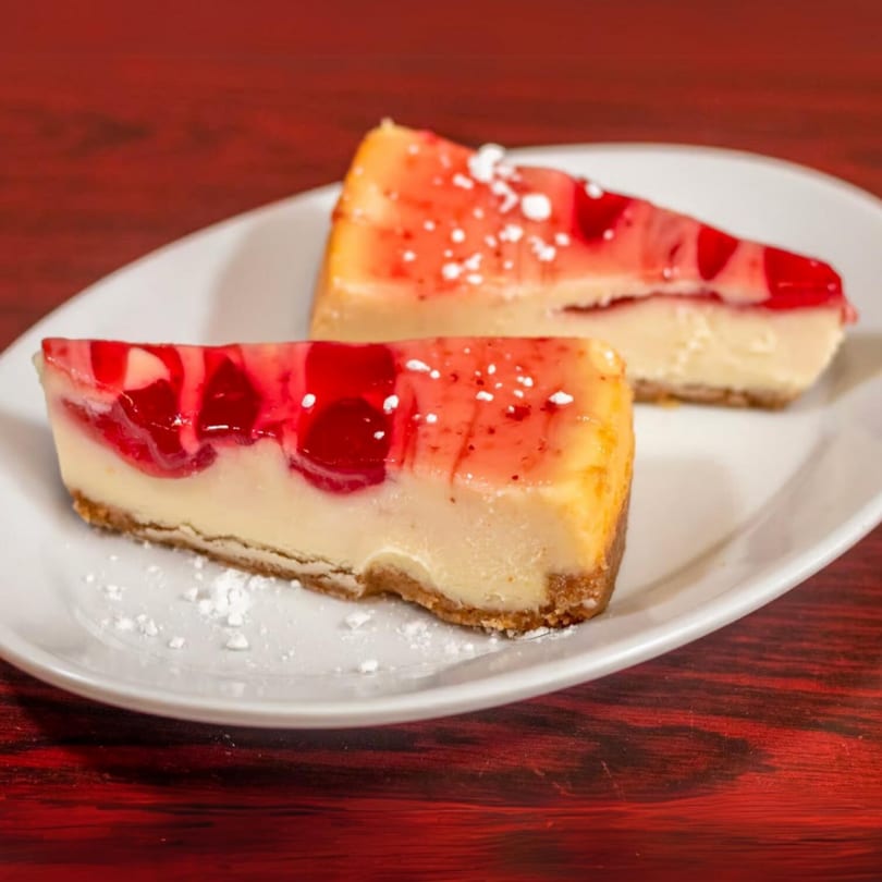 Two slices of cheesecake with red fruit topping and powdered sugar on a white plate.