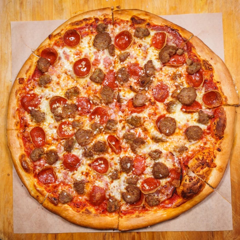 A whole, round pizza with pepperoni, sausage, and cheese, on a wooden surface.